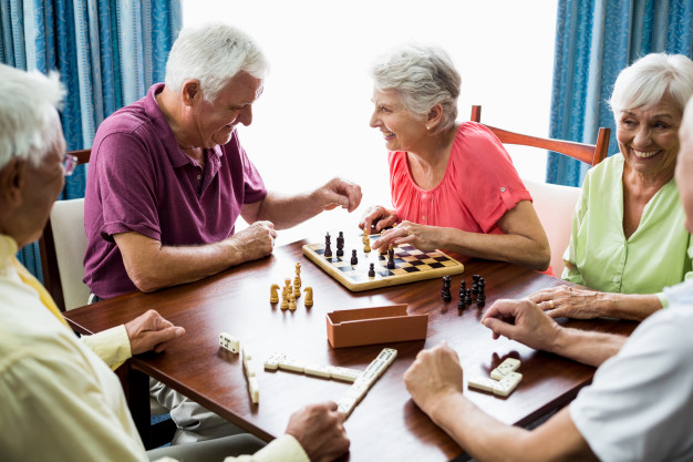 dois senhores de idade sentados em uma mesa do lado esquerdo de uma mesa enquanto há duas senhoras sentadas do lado direito, eles estão jogando xadrez e dominó, sorrindo